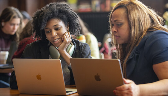 Two people looking at their laptops in a crowded room.